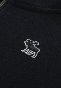 Brodert hvit og mørkegrå elglogo på svart teksturert stoff nær en sølvglidelås.