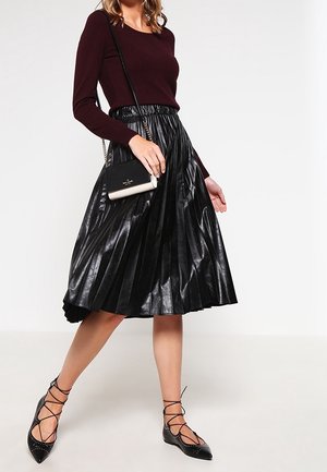 Femme portant un haut bordeaux à manches longues, une jupe midi plissée en cuir noir, des ballerines noires à lacets, et portant un petit sac bandoulière noir et blanc.