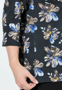 Bluse mit Blumenmuster auf schwarzem Hintergrund, mit blauen und braunen Blumen. Der Stoff ist weich, hat drei Viertelärmel und einen seitlichen Schlitz.