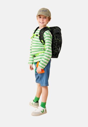 Schwarzer Rucksack mit grünen geometrischen Akzenten, grün-weiß gestreiftes Langarmshirt, blaue Jeansshorts, weiße Schuhe mit grünem Logo.