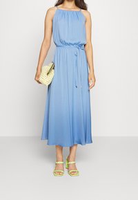 Hellblaues Maxikleid mit Halsband und gerafftem Oberteil, tailliert in der Taille. Kombiniert mit grünen Riemchen-Heels und einer gelben geflochtenen Clutch.