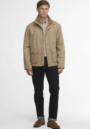 Man draagt beige jasje, lichtbruine shirt, donkere jeans met omslag aan de onderkant, grijze sokken en bruine loafers, staand tegen een witte achtergrond.