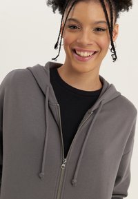 Felpa grigia con zip e cordini, realizzata in tessuto liscio, indossata sopra una maglietta nera a coste. Presenta una zip argentata e un fit casual.