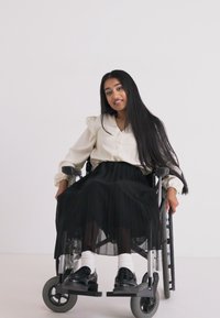 Jupe plissée noire avec une blouse bouton-pression crème, portée par une personne en fauteuil roulant. Tissus lisses, design minimaliste, se détache sur un fond neutre.