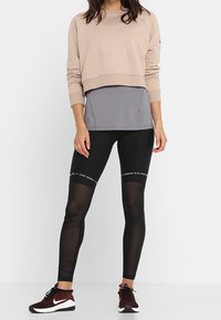 Beige kort sweatshirt med texturerad detalj, grå baslager och svarta leggings med genomskinliga paneler och vita textaccenter.