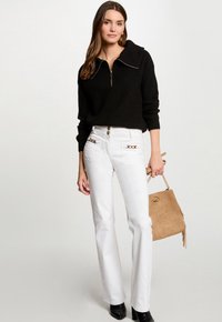 Black sweater with a zip collar, white flared jeans with gold hardware details, and a beige handbag with fringe accents. Black heeled boots.