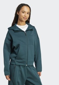 Tyrkysová mikina na zips vyrobená z mäkkej látky, s vysokým golierom, prednými vreckami a voľným strihom. Skombinovaná so zodpovedajúcimi nohavicami.