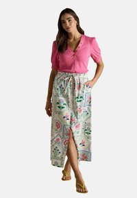 Frau in bluse mit kurzen Ärmeln in Pink, blumengemusterter Hochzeitsrock mit Schlitz an der Vorderseite und gelben Riemchensandalen.