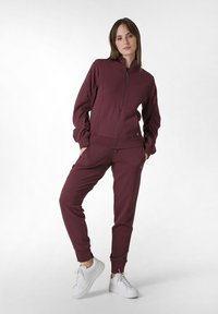 Bordeaux atletisk träningsdräkt med en zipjacka och matchande byxor. Inkluderar ribbade ärmslut, sidofickor och är ihop med vita sneakers.