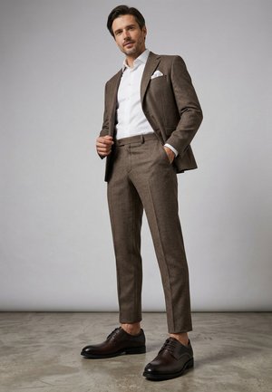 Mann trägt einen braunen strukturierten Anzug mit weißem Hemd und Einstecktuch, steht auf poliertem Betonboden vor einem schlichten grauen Hintergrund.