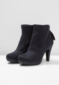 Botas de tobillo de ante negro con un tacón alto, cremallera lateral y borlas decorativas. Textura lisa y punta redondeada. Acento de plataforma brillante.