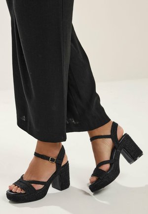 Voeten met zwarte geweven sandalen met hoge hakken en enkelbandjes, gecombineerd met zwarte brede broekspijpen op een lichte achtergrond.