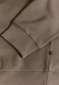 Sudadera marrón con tejido texturizado, presenta un puño acanalado, un logotipo sutil y líneas limpias. El diseño enfatiza la simplicidad y la comodidad.