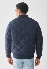 Marineblå quiltet bomberjakke med diamantmønster, ribbestrikkede mansjetter og kant, samt glidelås foran. Brukt over lyse denimjeans.