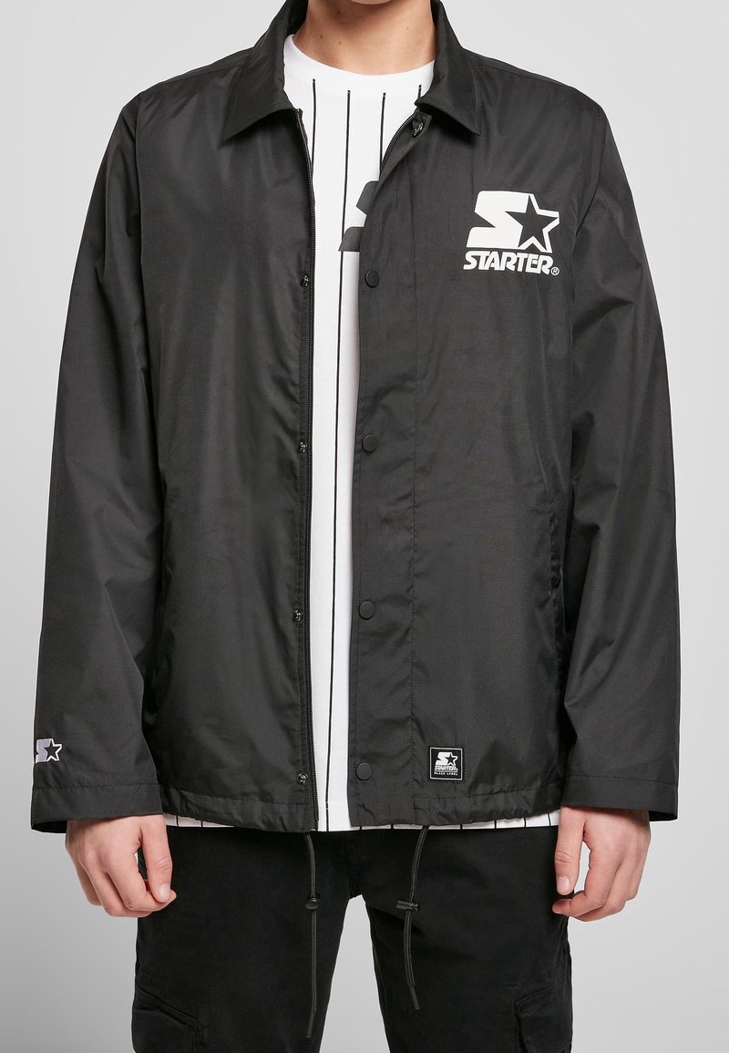 Personne portant une veste noire de la marque Starter par-dessus une chemise blanche à rayures verticales noires, debout devant un fond uni.