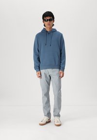 Felpa blu con cappuccio e tasca frontale, abbinata a jeans grigi chiari e sneakers bianche con suola beige. Occhiali da sole neri indossati.