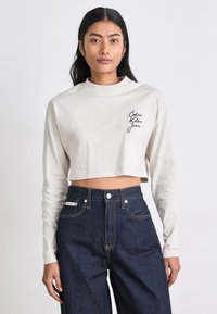 Top beige à manches longues coupé, avec un col rond, arborant le logo noir "Calvin Klein Jeans" sur le côté gauche. Porté avec un jean taille haute bleu foncé.
