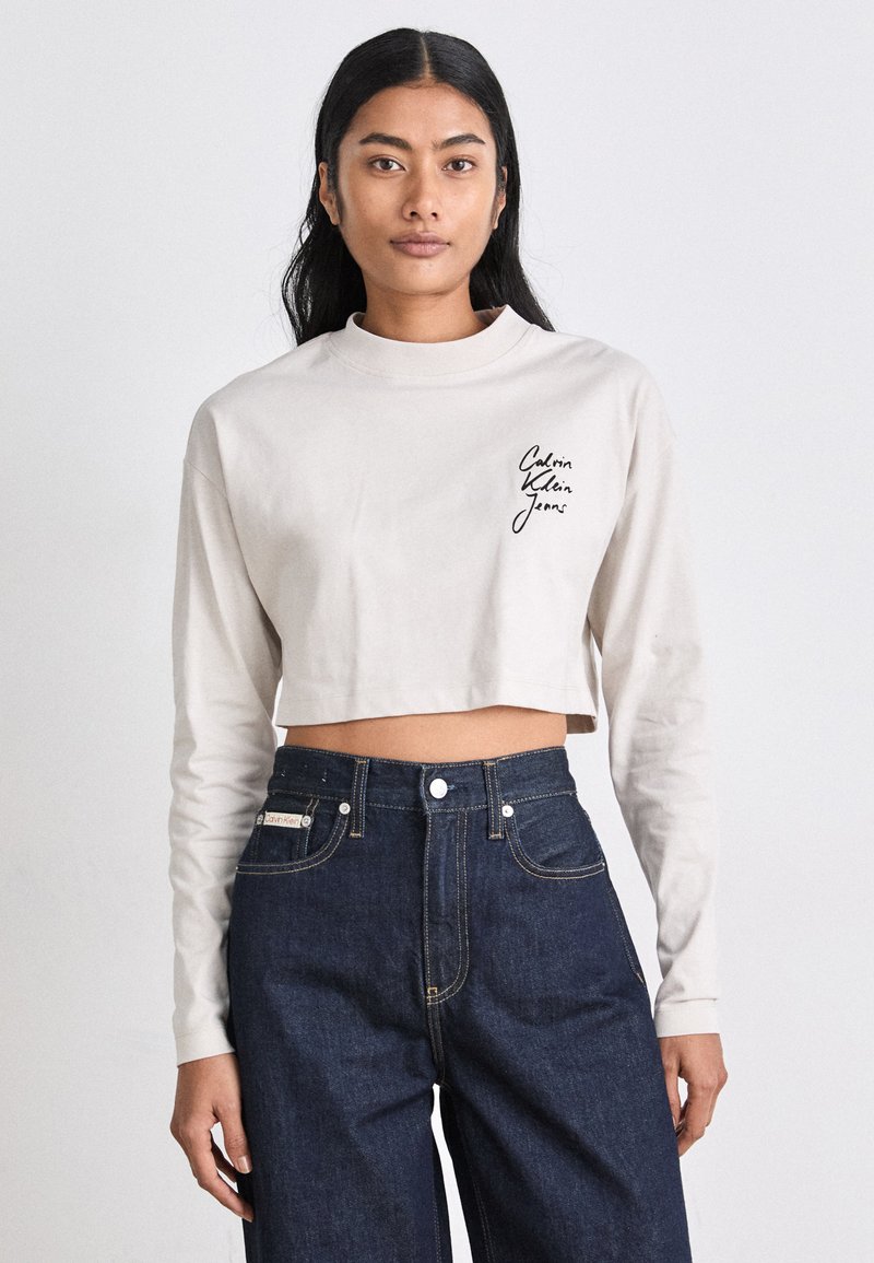 Top beige à manches longues coupé, avec un col rond, arborant le logo noir "Calvin Klein Jeans" sur le côté gauche. Porté avec un jean taille haute bleu foncé.