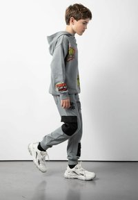 Szara bluza z kapturem z graficznymi nadrukami i tekstem, w połączeniu z pasującymi szarymi dresami z czarnymi akcentami. Białe sportowe buty dopełniają całość.