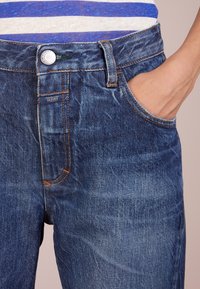 Tmavě modré džíny z denimu s mírným vyblednutím, mají přední kapsu, stříbrné zapínání na knoflík a oranžové švy.