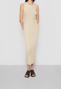 Beige žebrované pletené šaty bez rukávů s těsným střihem, sahajícími do poloviny lýtek. Spárováno s černými oversized sandály na klínu.