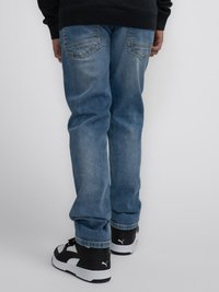 Jeans de mezclilla azul claro con corte recto y costuras visibles, combinados con zapatillas negras y blancas que tienen un logo en el lateral.