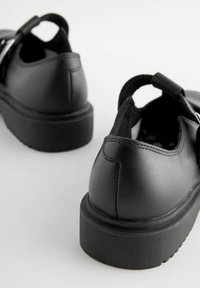 Chaussures en cuir noir avec un bout arrondi, une semelle en caoutchouc noir épaisse et un détail de sangle à l'arrière. Texture lisse avec des coutures visibles.
