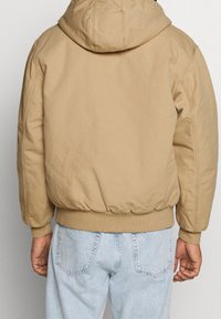 Beżowa kurtka z kapturem, z elastycznym paskiem w talii i mankietami, o pikowanej fakturze i widocznych szwach. Nosi ją nad jasnoniebieskimi dżinsami.