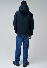 Veste matelassée bleu marine avec une grande capuche, finition texturée et poignets élastiques ; associée à un jean bleu ample et des bottes noires épaisses.