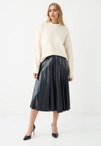 Maglione over-size di colore crema con polsini a coste, abbinato a una gonna midi plissettata in vinile nera e scarpe con tacco a punta nere.