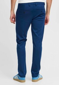 Moški v modrih oprijetih hlačah in svetlo modrih semiš športnih copatih z belimi črtami, ki stoji pred enobarvno belo ozadjem.