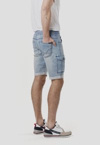 Pantaloncini cargo in denim azzurro chiaro con risvolti cuciti, vita elasticizzata, tasche laterali e dettagli di cucitura a contrasto. Indossati con sneakers.