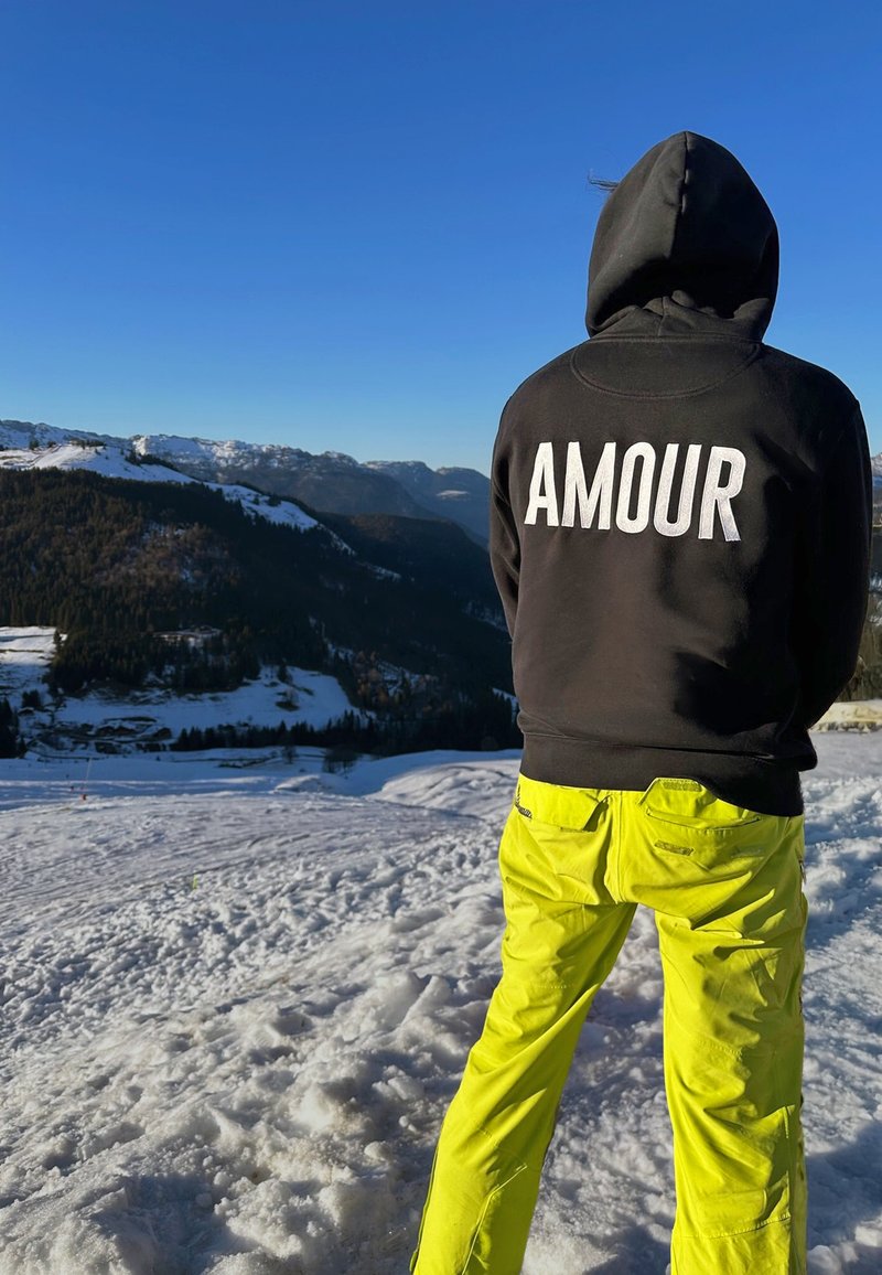 Črna kapuca z napisom "AMOUR" na hrbtu, v kombinaciji s svetlo rumenimi smučarskimi hlačami. Zunanja postavitev z snegom in gorami v ozadju.