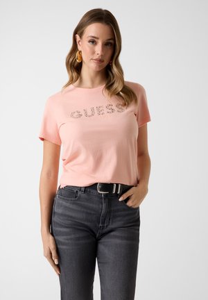 Femme aux cheveux ondulés portant un t-shirt rose Guess et un jean gris foncé taille haute avec une ceinture noire, posant avec une main dans la poche.