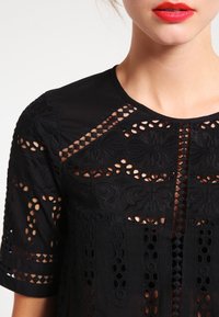 Svart topp med intrikata cut-out-mönster, texturerad broderi och en rundad halsringning. Korta ärmar med dekorativa detaljer.