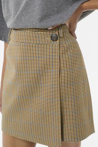 Jupe à carreaux jaune et marron avec un accent bleu. Dispose d'une fermeture à bouton unique à la taille et d'un design plissé sur un côté.