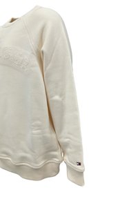 Felpa crema con logo di marca testurizzato, scollatura rotonda, orlo e polsini a costine, e una piccola etichetta logata colorata sulla manica.