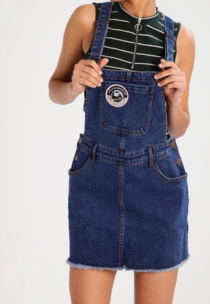 Femme portant un haut rayé sans manches et une jupe salopette en jean bleu, tenant les bretelles de la salopette avec les deux mains.