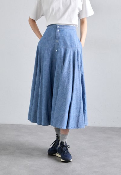 Hellblauer Denim-Rock mit ausgestelltem Design, hochgeschnitten, mit knopfbeschlägen hinten und seitlichen Taschen. Kombiniert mit marineblauen Sneakers und grauen Socken.