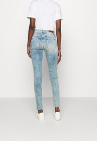 Jeans skinny de denim azul claro con un acabado desgastado, que cuentan con dos bolsillos traseros y una etiqueta de cuero en la cintura. Llevados con una camiseta blanca.