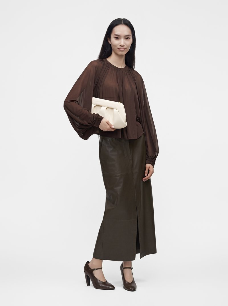 Femme portant un chemisier marron transparent, une jupe en cuir vert foncé, des talons marron, tenant une pochette crème, debout devant un fond uni.
