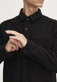 Camisa negra de botones con cuello, hecha de tejido texturizado. Presenta dos bolsillos delanteros y botones negros en la tapeta y los puños.