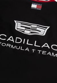 Black garment with white Cadillac logo and "Cadillac Formula 1 Team" text, featuring a Tommy Hilfiger label at the collar.