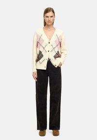 Kardigan w kolorze kremowym z dekoltem w serek, z diamentowym wzorem w kolorze różowym, szarym i bordowym, z zapięciem na guziki i prążkowanymi mankietami. Połączony z ciemnymi spodniami.