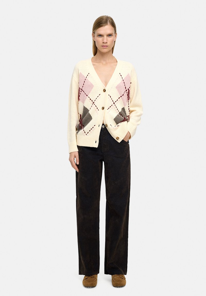 Kardigan w kolorze kremowym z dekoltem w serek, z diamentowym wzorem w kolorze różowym, szarym i bordowym, z zapięciem na guziki i prążkowanymi mankietami. Połączony z ciemnymi spodniami.