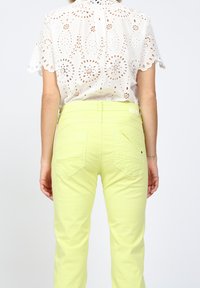 Ljusgula byxor med en slät textur och två bakfickor, tillsammans med en vit ögla spets-topp med vågformad nederkant och detaljerade utskärningar.