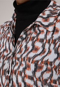 Chemise à boutons avec un motif ondulé en orange, noir et blanc. Col court, tissu texturé et un col roulé noir en dessous.
