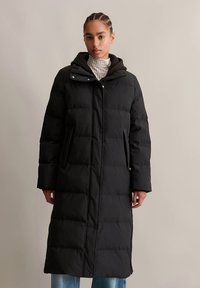 Γυναίκα ντυμένη με ένα μακρύ μαύρο καπιτονέ μπουφάν με κουκούλα, συνδυασμένο με ανοιχτόμπλε τζιν και καρό πουλόβερ με ψηλό γιακά.