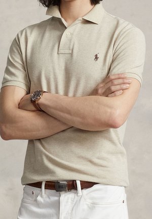 Homme portant un polo beige avec logo brodé, pantalon blanc, ceinture en cuir marron et montre, bras croisés sur un fond neutre.