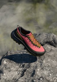Roza in črna La Sportiva pohodna čevlja z rumenimi vezalkami v zraku nad strnjeno sivo skalo na prostem.